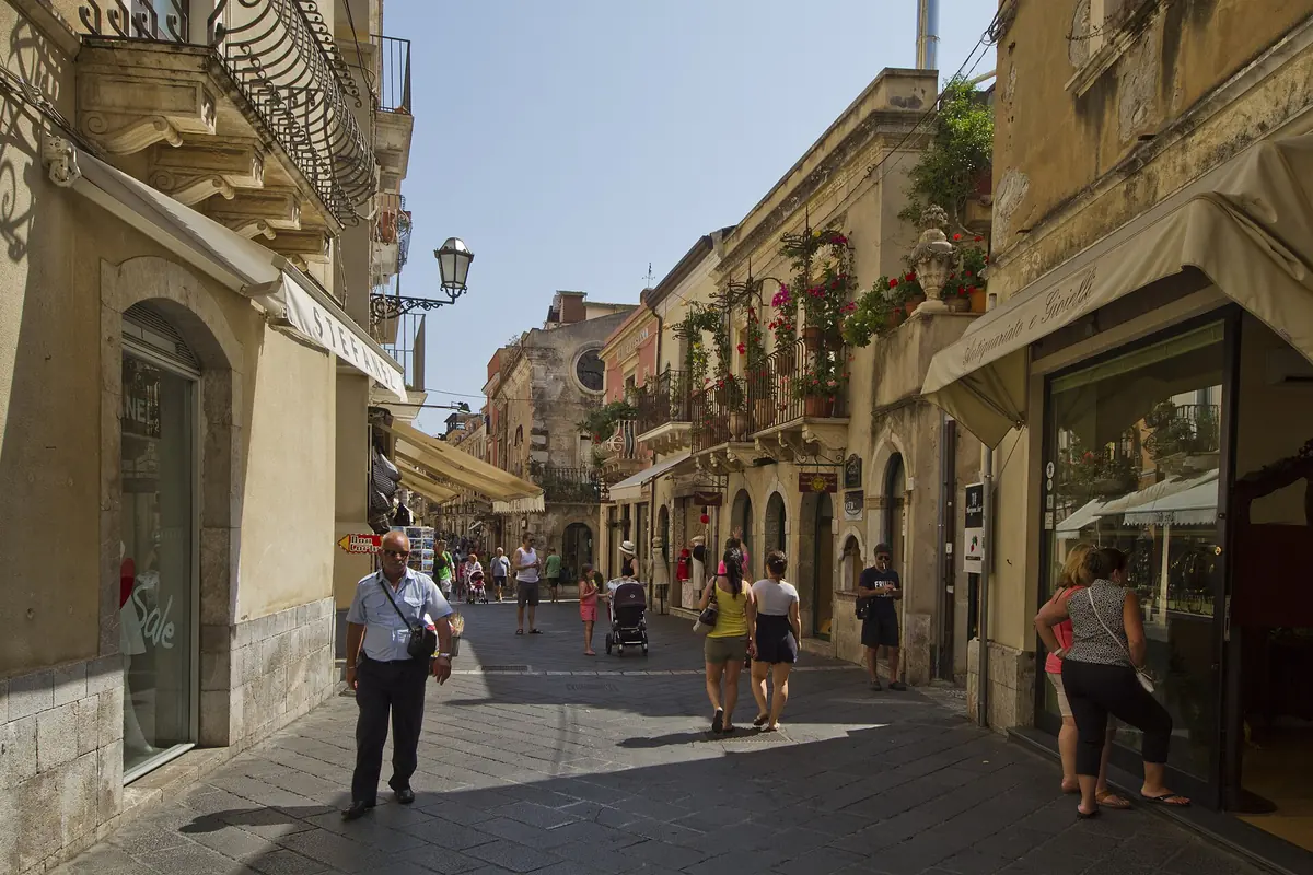 Corso Umberto in Taormina by day, a narrow pedestrian street between old stone buildings with shop awnings and iron balconies, and a few people walking.