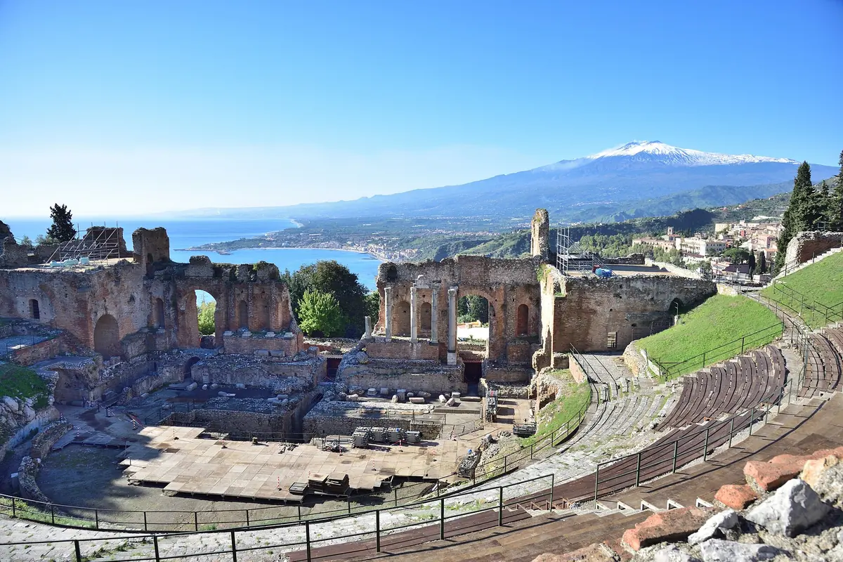 The ruined stage wall and curved seating of Taormina's ancient theatre on a clear day, with the snow-capped cone of Mount Etna and the sea in the distance.