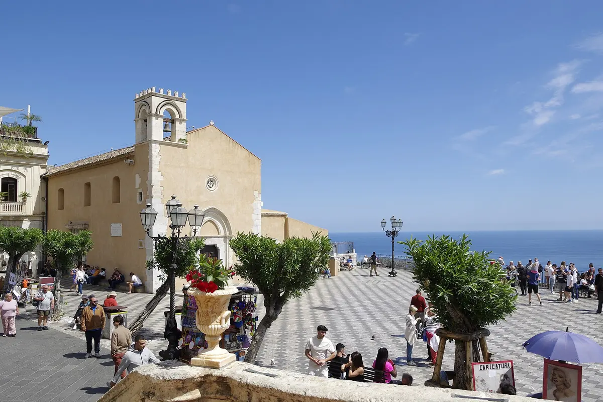 Piazza IX Aprile in Taormina by day, with a chequered stone floor, trees in pots, the church of San Giuseppe and a strip of blue sea beyond the terrace wall.