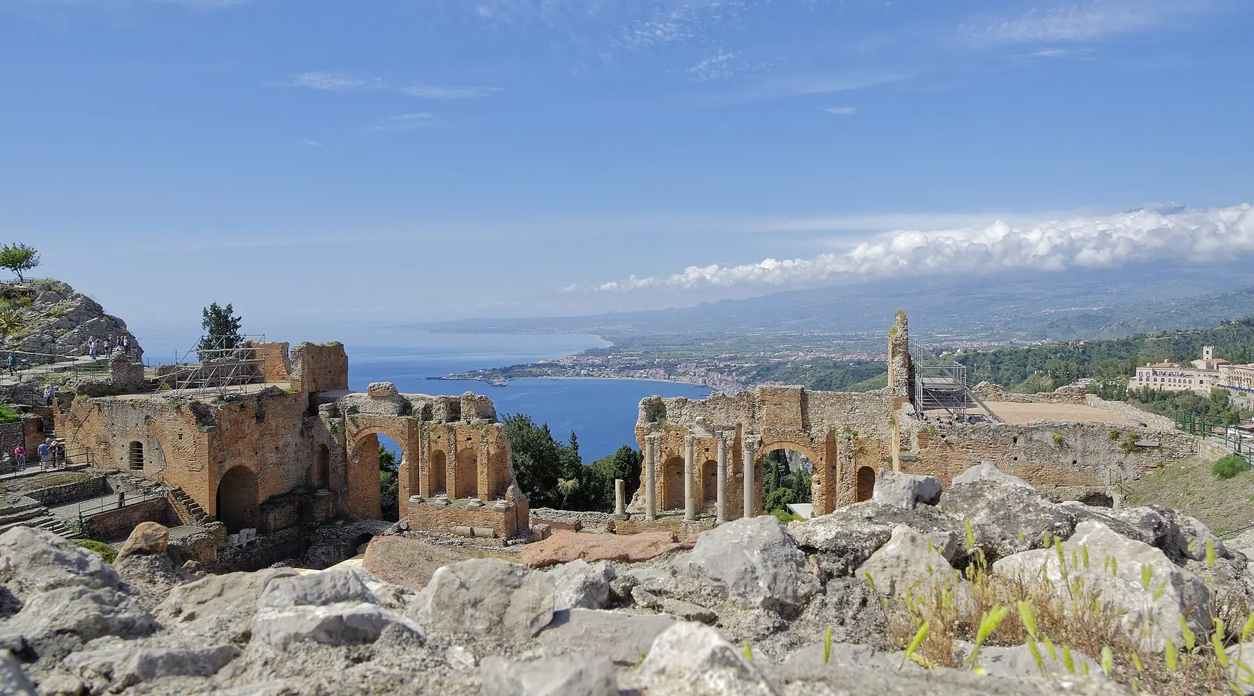 The ruined arches of Taormina's ancient theatre seen across rough rock, with the coastline and a long cloud bank on the horizon.