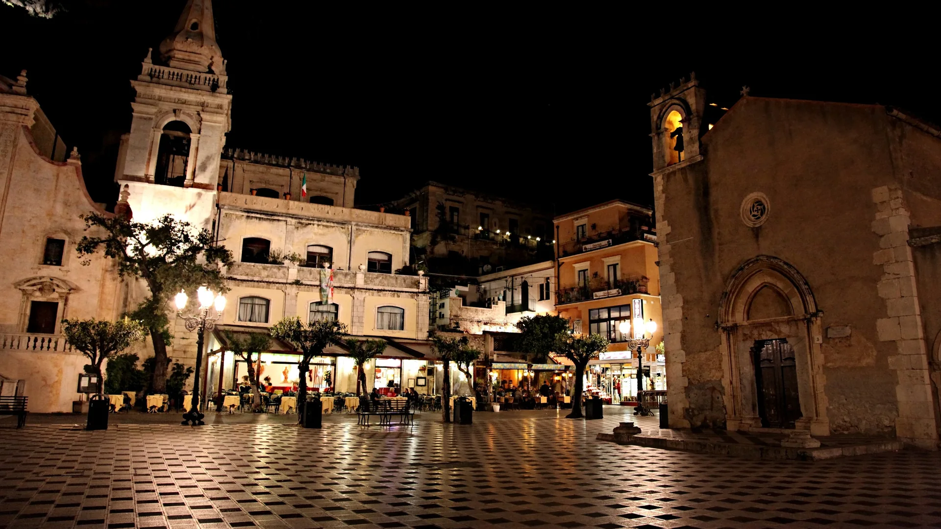 Piazza IX Aprile in Taormina at night, a chequered stone floor lit by street lamps, with the bell tower of the church of San Giuseppe on the left and a small stone church with a Gothic doorway on the right.